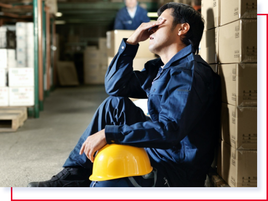 Imagen de un trabajador con un overol de trabajo azul y un casco amarillo sobre su pierna izquierda. Se encuentra exhausto y sentado. Se frota los ojos con su mano derecha.